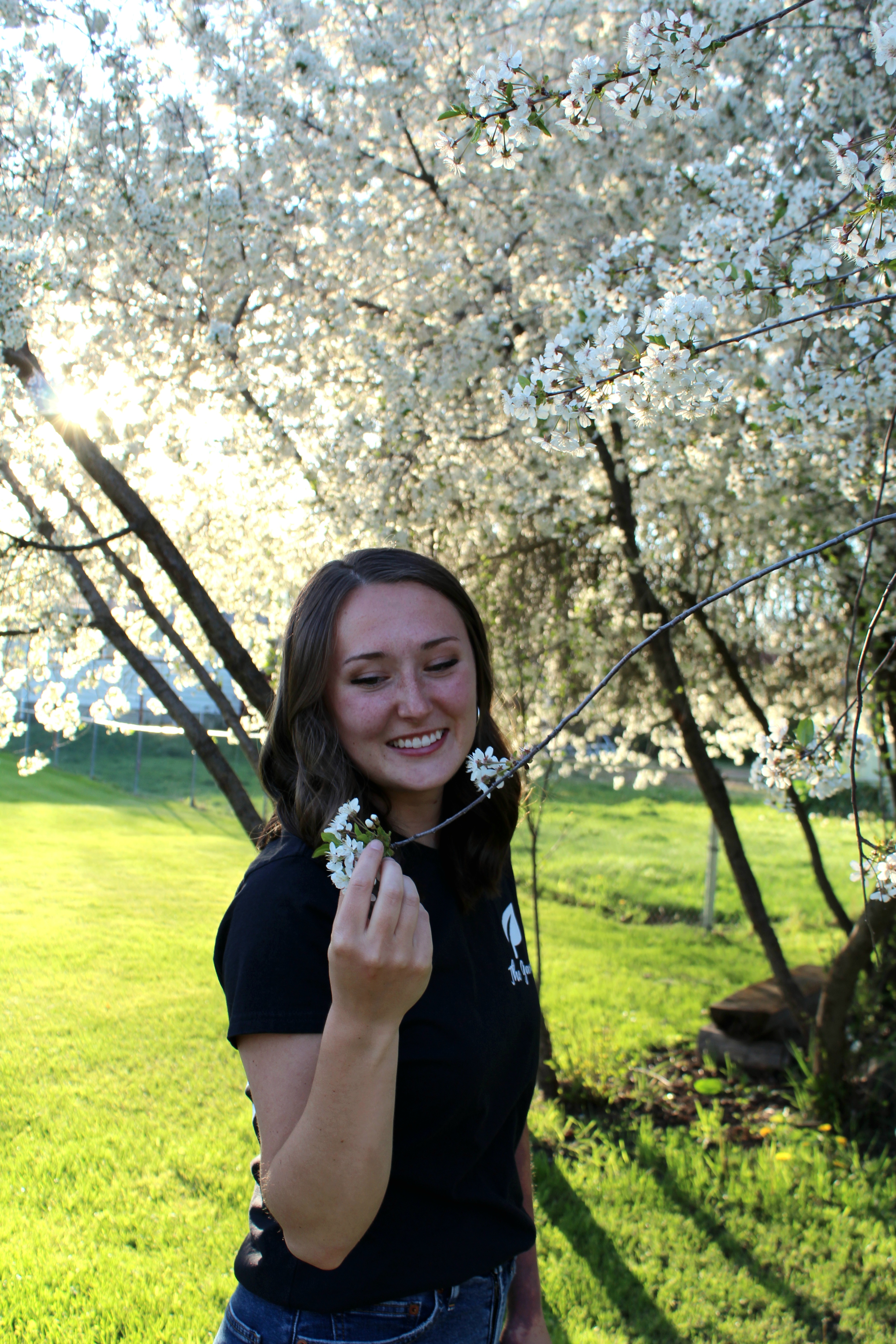 Gracie has shoulder length brown hair and is wearing a black t-shirt. She is standing underneath a tree with white flower buds.